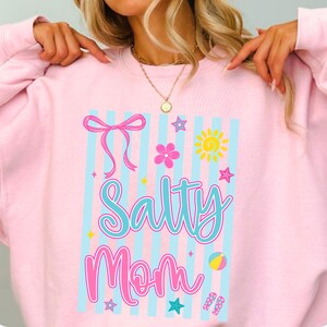 Pu&ograve; includere: Felpa rosa chiaro con la scritta "Salty Mom" in turchese. Il design include un fiocco rosa, strisce blu, un sole giallo e grafiche a tema spiaggia. La felpa &egrave; in stile casual.