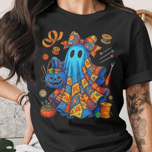 May include: Black t-shirt featuring a blue ghost with a patchwork quilt design. The ghost is holding a jack-o'-lantern basket and surrounded by sewing notions like spools of thread, needles, and buttons. A bow adorns the ghost's head.