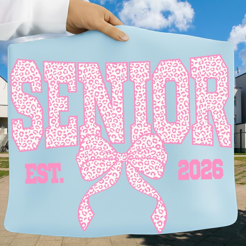 Puede incluir: Un letrero azul claro con la palabra "SENIOR" en letras rosas con estampado de leopardo. Debajo, un lazo rosa y el texto "EST. 2026". El letrero es sostenido por una persona.
