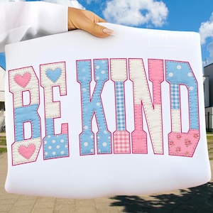 May include: A white tote bag displaying the words "BE KIND" in a patchwork design. The letters are in blue, pink, and white with polka dots, gingham, and floral patterns. The bag is held up against a blue sky.
