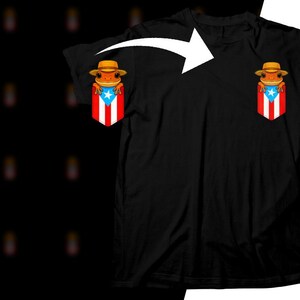 K&ouml;nnte beinhalten: Schwarzes T-Shirt mit einer Cartoon-Frosch, der einen Hut tr&auml;gt, in einer Tasche mit dem Design der puerto-ricanischen Flagge. Der Frosch ist orange und der Hut ist hellbraun. Das Flaggen-Design umfasst rote und wei&szlig;e Streifen, ein blaues Dreieck und einen wei&szlig;en Stern.