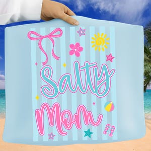 Pu&ograve; includere: Un telo mare azzurro chiaro con strisce verticali e la scritta "Salty Mom" in un carattere giocoso. L'asciugamano presenta grafiche a tema spiaggia rosa e gialle, tra cui un fiocco, stelle, un fiore, un sole, un pallone da spiaggia e infradito.