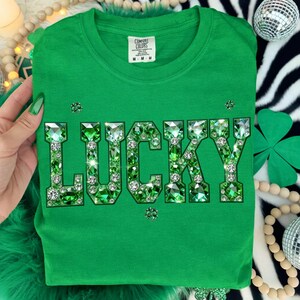 May include: A green t-shirt with the word "LUCKY" spelled out in large, sparkling green and clear gemstones. The shirt is a Comfort Colors brand and is displayed on a zebra-print background with St. Patrick's Day decorations.