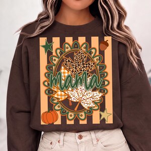 May include: A dark brown long-sleeve sweatshirt featuring a central graphic with the word mama in green cursive script. The design includes a circular frame, maple leaves with leopard and plaid patterns, a pumpkin, an acorn, and two stars.