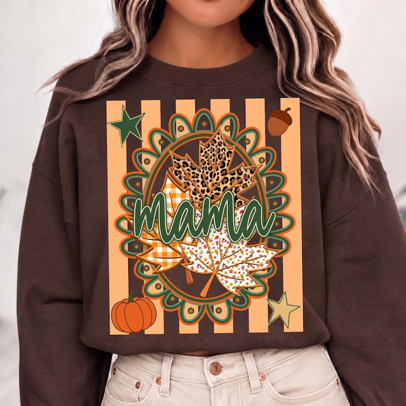 May include: A dark brown long-sleeve sweatshirt featuring a central graphic with the word mama in green cursive script. The design includes a circular frame, maple leaves with leopard and plaid patterns, a pumpkin, an acorn, and two stars.