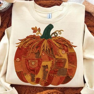 May include: A cream-colored crewneck sweatshirt featuring a large, central pumpkin graphic. The pumpkin is constructed from various patches of orange, brown, and yellow fabric with frayed edges, accented by small brown buttons and a dark green stem.