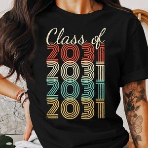 Puede incluir: Una camiseta negra con el texto "Class of 2031" impreso en un estilo retro con rayas de colores.