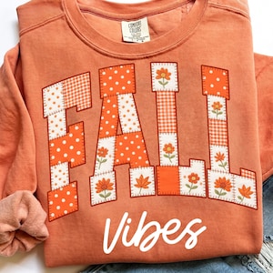 Op de afbeelding: Een terracotta-kleurige sweater met ronde hals en het woord FALL in grote blokletters gevuld met een patchworkpatroon van oranje en witte stippen, ruitjes en bloemmotieven, met het woord Vibes eronder geschreven in een wit cursief lettertype.