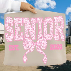 Puede incluir: Un bolso tote beige con la palabra "SENIOR" en letras grandes rosas con estampado de leopardo. Debajo de la palabra "SENIOR" hay un lazo rosa. El texto "EST. 2026" est&aacute; impreso debajo del lazo.