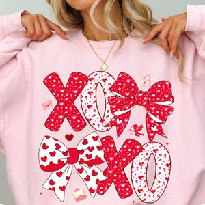 May include: Pink sweatshirt with a large XOXO design in red and white. The letters are decorated with hearts and bows, with small illustrations of envelopes, a lock, and Cupid. A Valentine's Day themed top.