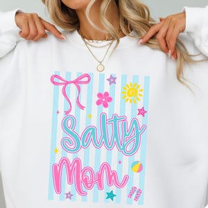Pu&ograve; includere: Felpa bianca con una grafica colorata che dice "Salty Mom". Il design include un fiocco rosa, un fiore, un sole, stelle, un pallone da spiaggia e infradito. Lo sfondo ha strisce blu e bianche.