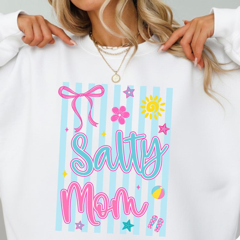 Pu&ograve; includere: Felpa bianca con una grafica colorata che dice "Salty Mom". Il design include un fiocco rosa, un fiore, un sole, stelle, un pallone da spiaggia e infradito. Lo sfondo ha strisce blu e bianche.