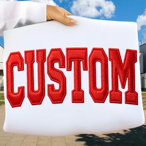 May include: A white pillow with the word "CUSTOM" embroidered in bold red letters. The letters have a collegiate style, with a thick, textured appearance. The pillow is held up against a blue sky background.