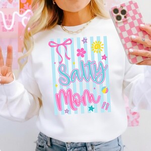 Pu&ograve; includere: Felpa bianca con grafica colorata con la scritta "Salty Mom". Il design include un fiocco rosa, fiori, stelle, un sole, un pallone da spiaggia e infradito. Lo sfondo presenta strisce verticali azzurre.