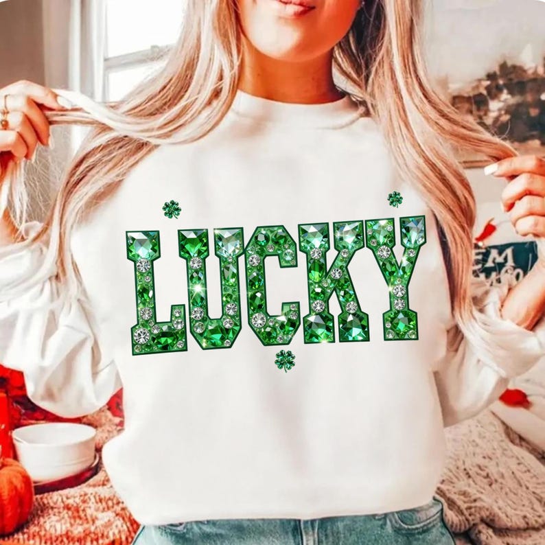 May include: White sweatshirt with the word "LUCKY" in large, sparkling green letters. The letters are embellished with green gems and small shamrock accents, creating a festive and eye-catching design.