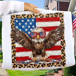 May include: A white pillow featuring an American flag design with a bald eagle wearing heart-shaped sunglasses. The eagle has outstretched wings, and the pillow has a leopard print border. The flag has red and white stripes and blue stars.