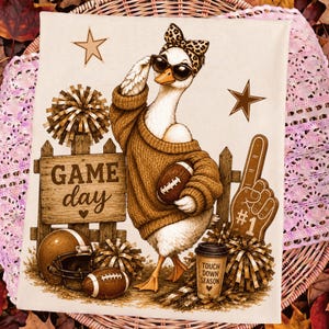 May include: A white fabric towel featuring a cartoon duck wearing a leopard print bow and sunglasses, a brown sweater, and holding a football. The design includes a wooden fence, a football helmet, pom-poms, a foam finger, and text reading GAME day.
