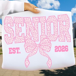 Puede incluir: Cartel blanco con texto rosa y diseño de lazo. La palabra "SENIOR" se muestra prominentemente, con un estampado de leopardo. Debajo, un lazo rosa. También se ve el texto "EST. 2026".