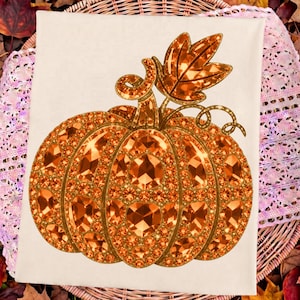 May include: A rectangular off-white fabric towel featuring a central graphic of a pumpkin composed of numerous orange and gold faceted gemstones. The pumpkin design includes a stem and a single leaf, all rendered with a sparkling, jewel-like texture.
