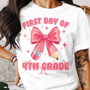 May include: White t-shirt with a pink graphic featuring a bow-shaped pencil and the text "FIRST DAY OF 4TH GRADE." The design includes pink stars and a bow at the bottom, perfect for back-to-school.