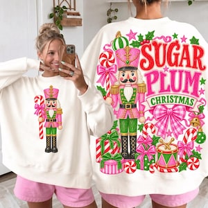 Puede incluir: Una sudadera blanca de manga larga con un gráfico de un cascanueces rosa y verde sosteniendo un bastón de caramelo en el frente, y un estampado grande en la espalda con un cascanueces, bastones de caramelo, regalos y el texto Sugar Plum Christmas.