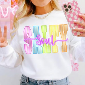 May include: White sweatshirt with the text "Salty Soul" in a colorful, striped design. The word "Salty" is in pink, blue, yellow, and orange. The word "Soul" is in a cursive, purple font.