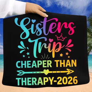 May include: Black beach towel with the text "Sisters Trip CHEAPER THAN THERAPY - 2026" in a colorful, gradient font. The design includes hearts, stars, and an arrow. The towel is held up against a beach backdrop.