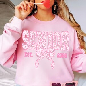 Puede incluir: Sudadera rosa con la palabra "SENIOR" en estampado de leopardo, un lazo y el texto "EST. 2026". La sudadera es de color rosa liso.