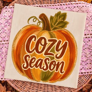 Könnte beinhalten: Eine quadratische Leinwand mit einem gemalten orangefarbenen und gelben Kürbis mit braunem Stiel und grünem Blatt. Der Kürbis ist mit goldenem Glitzer umrandet. Der Text "COZY SEASON" steht in großen, dunkelbraunen Schreibschriftbuchstaben in der Mitte.