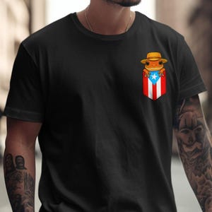K&ouml;nnte beinhalten: Schwarzes T-Shirt mit einem Taschenmotiv, das die puerto-ricanische Flagge und einen Cartoon-Frosch mit Hut zeigt. Der Frosch ist orange und der Hut ist hellbraun. Das T-Shirt hat eine einfarbige schwarze Farbe.