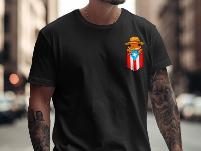 K&ouml;nnte beinhalten: Schwarzes T-Shirt mit einem Taschenmotiv, das die puerto-ricanische Flagge und einen Cartoon-Frosch mit Hut zeigt. Der Frosch ist orange und der Hut ist hellbraun. Das T-Shirt hat eine einfarbige schwarze Farbe.