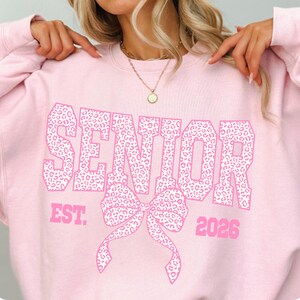 Puede incluir: Sudadera rosa claro con la palabra "SENIOR" en letras grandes con estampado de leopardo. Debajo, un lazo con el mismo patr&oacute;n y el texto "EST. 2026". La sudadera es de cuello redondo.