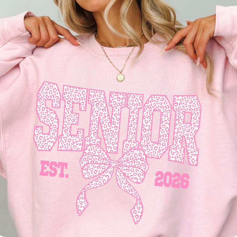 Puede incluir: Sudadera rosa claro con la palabra "SENIOR" en letras grandes con estampado de leopardo. Debajo, un lazo con el mismo patr&oacute;n y el texto "EST. 2026". La sudadera es de cuello redondo.