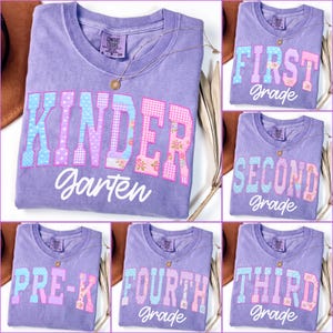 May include: A purple short-sleeved t-shirt featuring a variety of school grade levels printed in large, block-style letters with mixed patterns including polka dots, gingham, and floral prints, accompanied by the word grade in white script.
