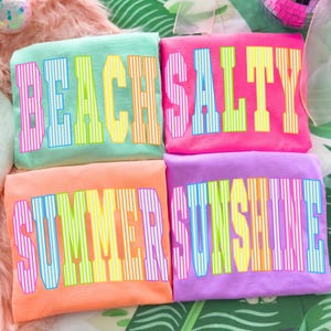 May include: Four colorful t-shirts with bold text. The shirts are in shades of mint green, pink, orange, and purple. Each shirt has a different word: "BEACH", "SALTY", "SUMMER", and "SUNSHINE" in a striped font.