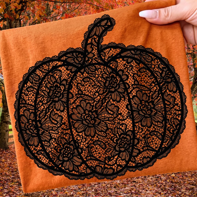 May include: A burnt orange t-shirt featuring a large, black graphic of a pumpkin. The pumpkin design is composed of intricate, detailed black lace patterns with floral motifs, creating a textured, decorative appearance on the fabric.