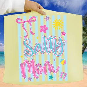 Pu&ograve; includere: Un telo da mare giallo chiaro con strisce blu e bianche. Il telo presenta le parole "Salty Mom" in corsivo turchese e rosa, insieme a grafiche a tema spiaggia come un sole, stelle e un pallone da spiaggia.