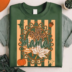 May include: A dark green crewneck sweatshirt featuring a central graphic with the word mama in cursive script. The design includes a scalloped oval frame, maple leaves with leopard and plaid patterns, a pumpkin, an acorn, and two stars.