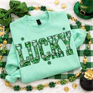 May include: A mint green sweatshirt with the word "LUCKY" in large, sparkling green gem lettering. The design includes shamrocks and is surrounded by St. Patrick's Day themed decorations, including a green bow, gold coins, and a leprechaun hat.