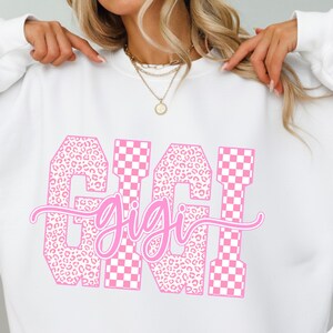 May include: White sweatshirt with a pink graphic that says "GIGI". The letters are a mix of leopard print and pink and white checkered patterns. The word "gigi" is written in a cursive font.