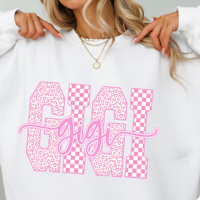 May include: White sweatshirt with a pink graphic that says "GIGI". The letters are a mix of leopard print and pink and white checkered patterns. The word "gigi" is written in a cursive font.