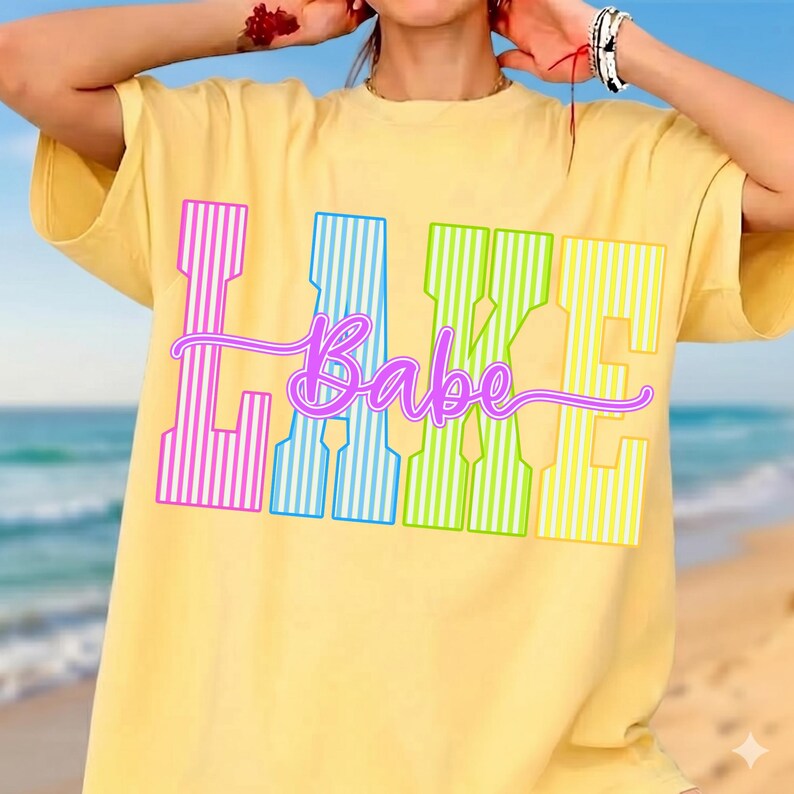 May include: Yellow t-shirt with the word "Lake Babe" in a colorful, striped design. The word "Lake" is in pink, blue, and green stripes, and "Babe" is in a cursive font. The shirt has short sleeves.