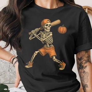 Puede incluir: Camiseta negra con un esqueleto con pantalones cortos naranjas, una gorra de béisbol y zapatillas, balanceando un bate hacia una calabaza. El diseño tiene un tema de Halloween.