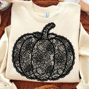 May include: A cream-colored crewneck sweatshirt featuring a large, centered graphic of a pumpkin. The pumpkin design is rendered in a black, intricate lace pattern with detailed floral motifs throughout the shape, including a curved stem.