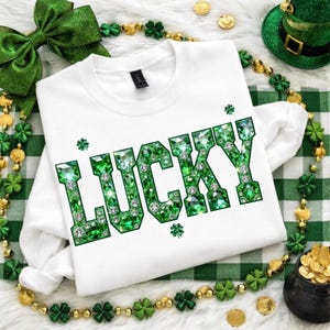 May include: White crew neck sweatshirt with the word "LUCKY" in large, sparkling green gem lettering. The design includes shamrocks and is surrounded by St. Patrick's Day themed decorations, including a green bow and a pot of gold.