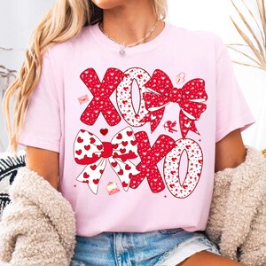 May include: A light pink t-shirt featuring the letters "XOXO" in a red bow design with white hearts. The design includes small red hearts, envelopes, and a cupid illustration. The shirt is a Valentine's Day theme.