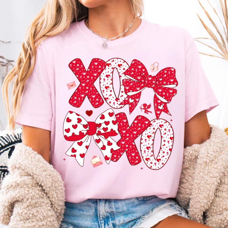 May include: A light pink t-shirt featuring the letters "XOXO" in a red bow design with white hearts. The design includes small red hearts, envelopes, and a cupid illustration. The shirt is a Valentine's Day theme.