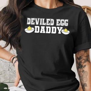 Può includere: T-shirt nero con la scritta bianca "DEVILED EGG DADDY" e grafiche di uova alla diavola. Una t-shirt casual per l'uso quotidiano.