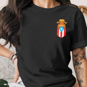 K&ouml;nnte beinhalten: Schwarzes T-Shirt mit einer Frosch-Illustration, der einen Hut tr&auml;gt, in einer Tasche mit der Flagge von Puerto Rico. Der Frosch ist orange und der Hut ist hellbraun. Die Tasche hat rote und wei&szlig;e Streifen und einen blauen Stern.