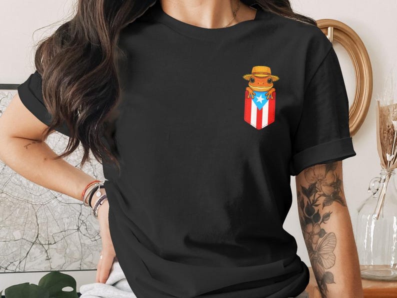 K&ouml;nnte beinhalten: Schwarzes T-Shirt mit einer Frosch-Illustration, der einen Hut tr&auml;gt, in einer Tasche mit der Flagge von Puerto Rico. Der Frosch ist orange und der Hut ist hellbraun. Die Tasche hat rote und wei&szlig;e Streifen und einen blauen Stern.